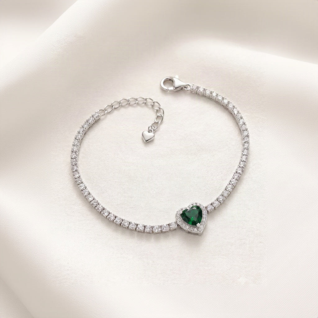 A silver tennis-style bracelet with a heart-shaped green gemstone centerpiece, displayed on a soft cream satin background.