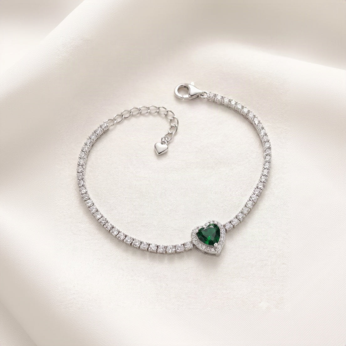 A silver tennis-style bracelet with a heart-shaped green gemstone centerpiece, displayed on a soft cream satin background.