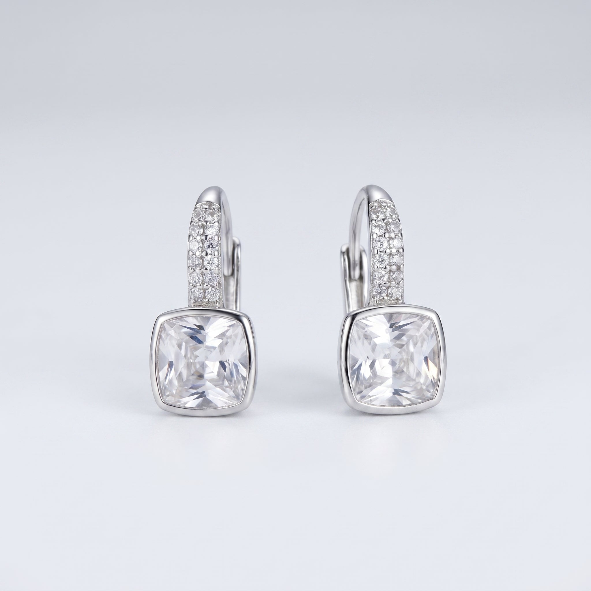 A pair of silver leverback earrings featuring a large cushion-cut clear gemstone in a bezel setting, with a pavé band of small clear stones above it, shown on a light gray gradient background.