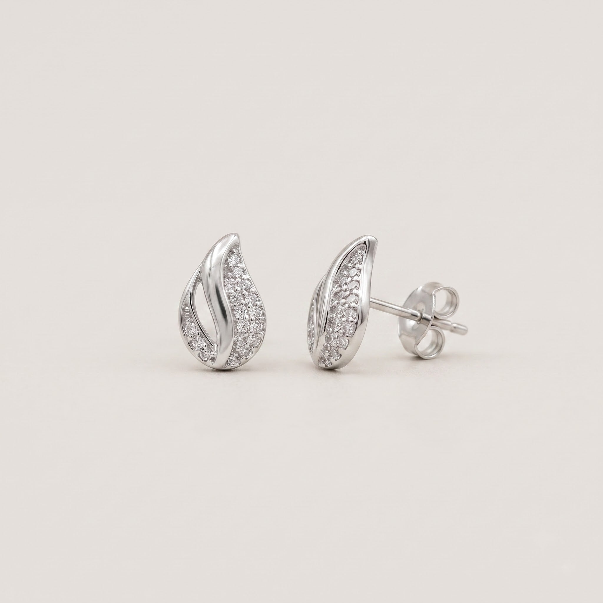 A pair of silver teardrop-shaped stud earrings paved with small clear stones, shown from both front and side angles on a light beige background.