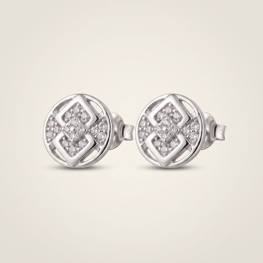 A pair of round silver stud earrings with a geometric interlocking pattern set with small clear pavé stones, shown on a light beige background.