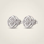 A pair of round silver stud earrings with a geometric interlocking pattern set with small clear pavé stones, shown on a light beige background.