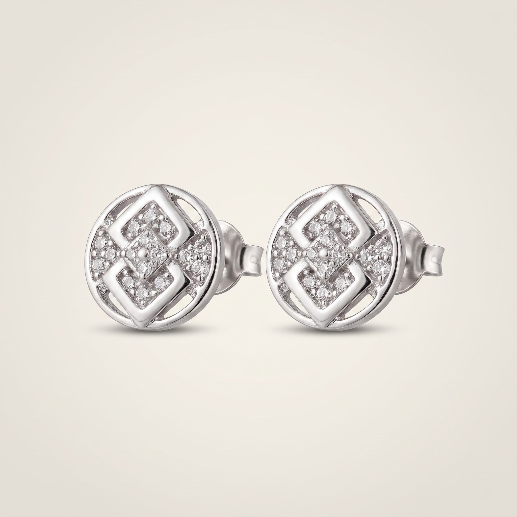 A pair of round silver stud earrings with a geometric interlocking pattern set with small clear pavé stones, shown on a light beige background.