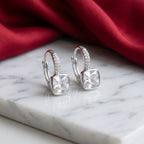 A pair of silver huggie hoop earrings with square clear gemstones and small pavé-set accents, displayed on a white marble slab with a red fabric backdrop.