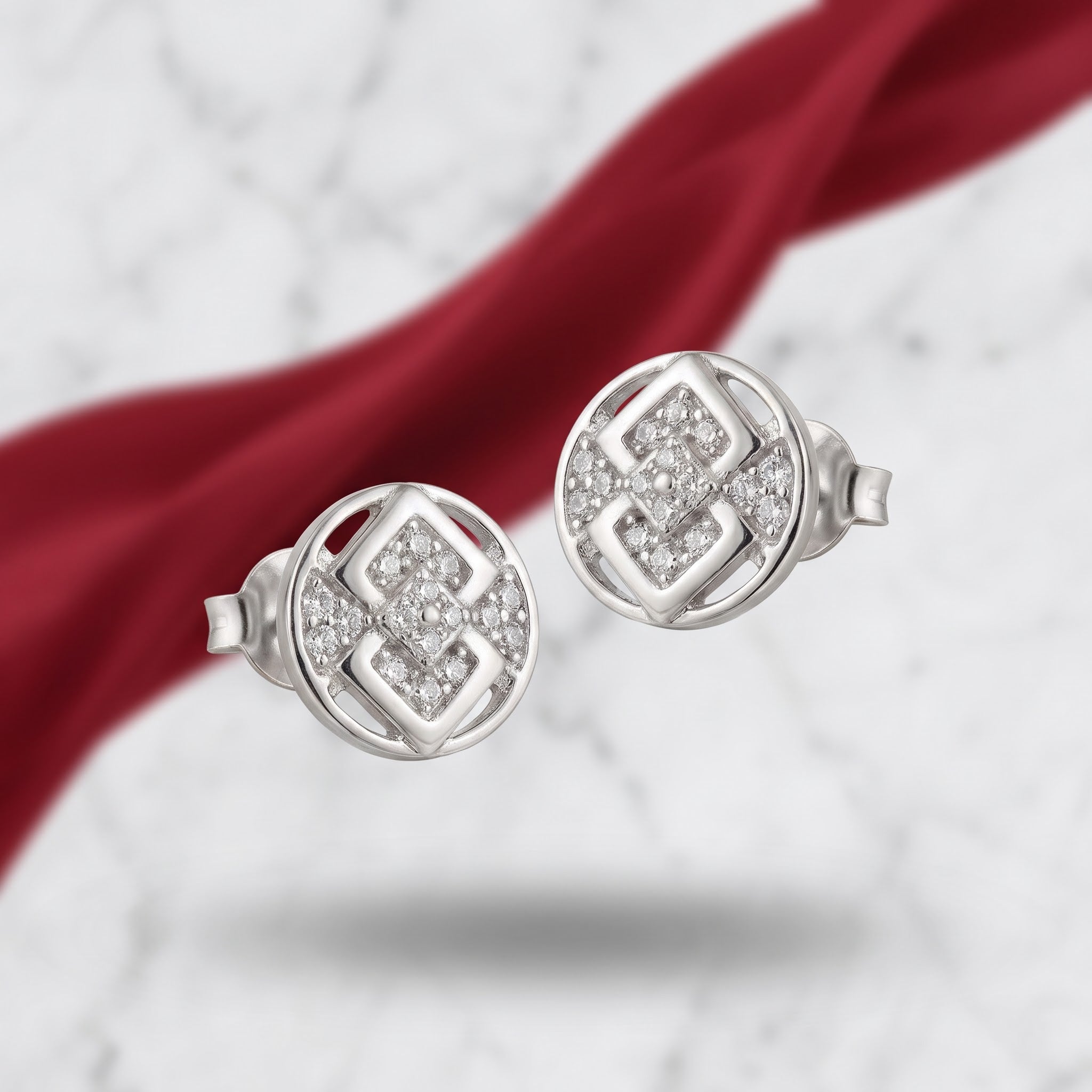 A pair of round silver stud earrings with pavé-set clear stones and a geometric openwork design, displayed on a white marble surface with a draped red ribbon.