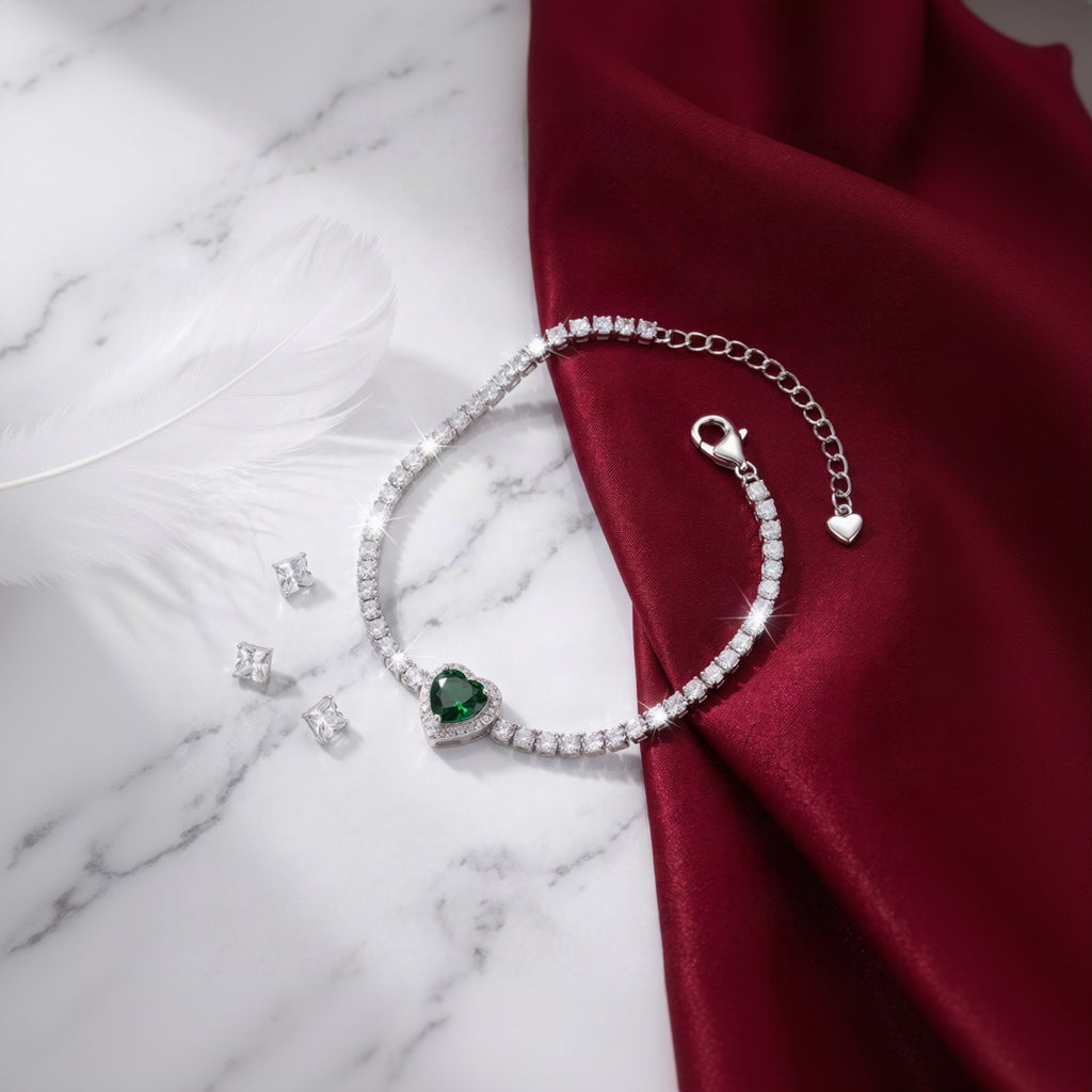 A silver-toned tennis bracelet with a heart-shaped green gemstone centerpiece, displayed on a white marble surface next to a draped dark red satin fabric.