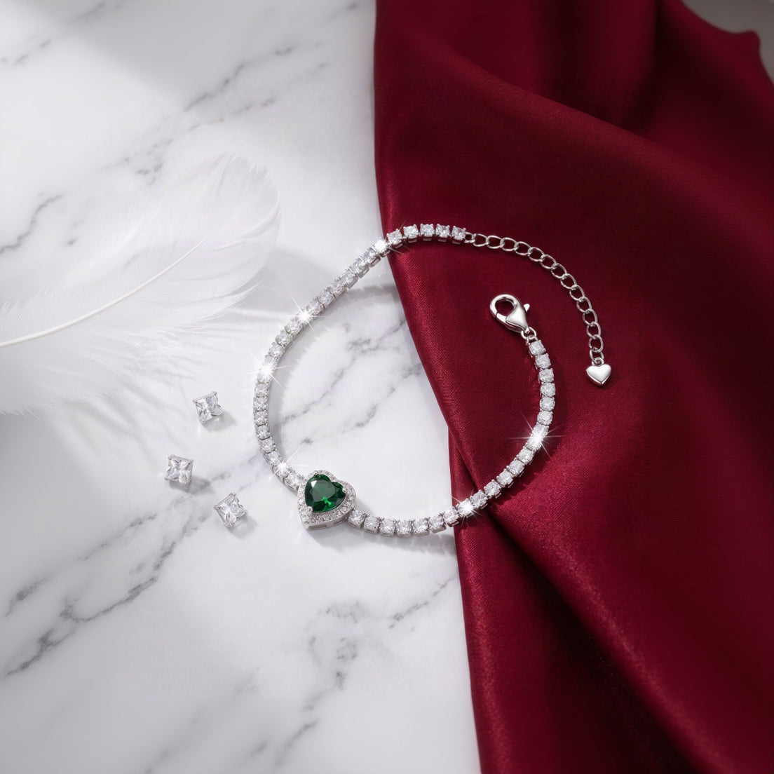 A silver-toned tennis bracelet with a heart-shaped green gemstone centerpiece, displayed on a white marble surface next to a draped dark red satin fabric.