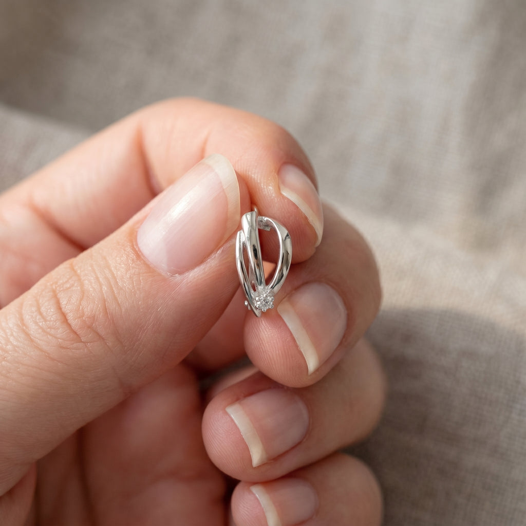 A close-up of a hand holding a small silver huggie hoop earring with a twisted ribbon-like design.