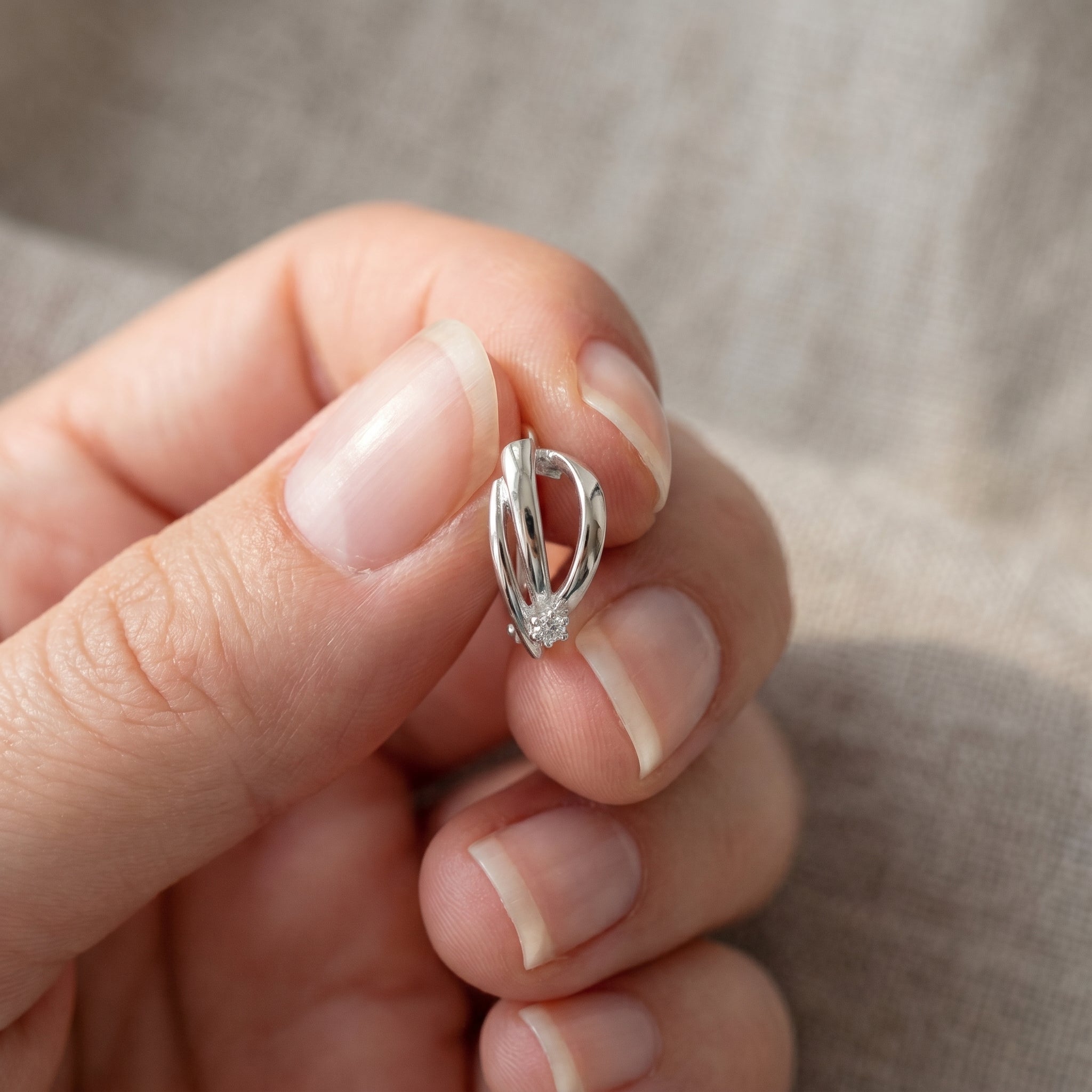 A close-up of a hand holding a small silver huggie hoop earring with a twisted ribbon-like design.