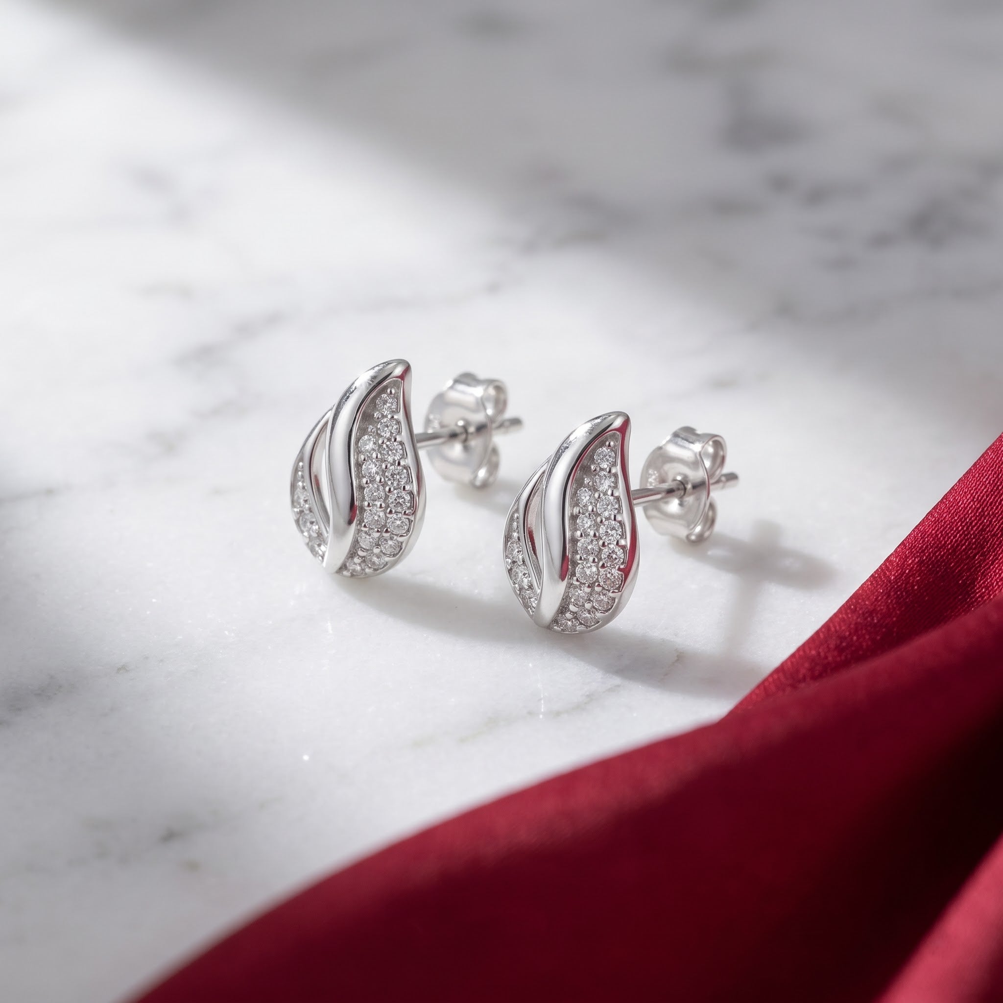 A pair of silver leaf-shaped stud earrings paved with small clear stones, displayed on a white marble surface next to a draped red fabric.