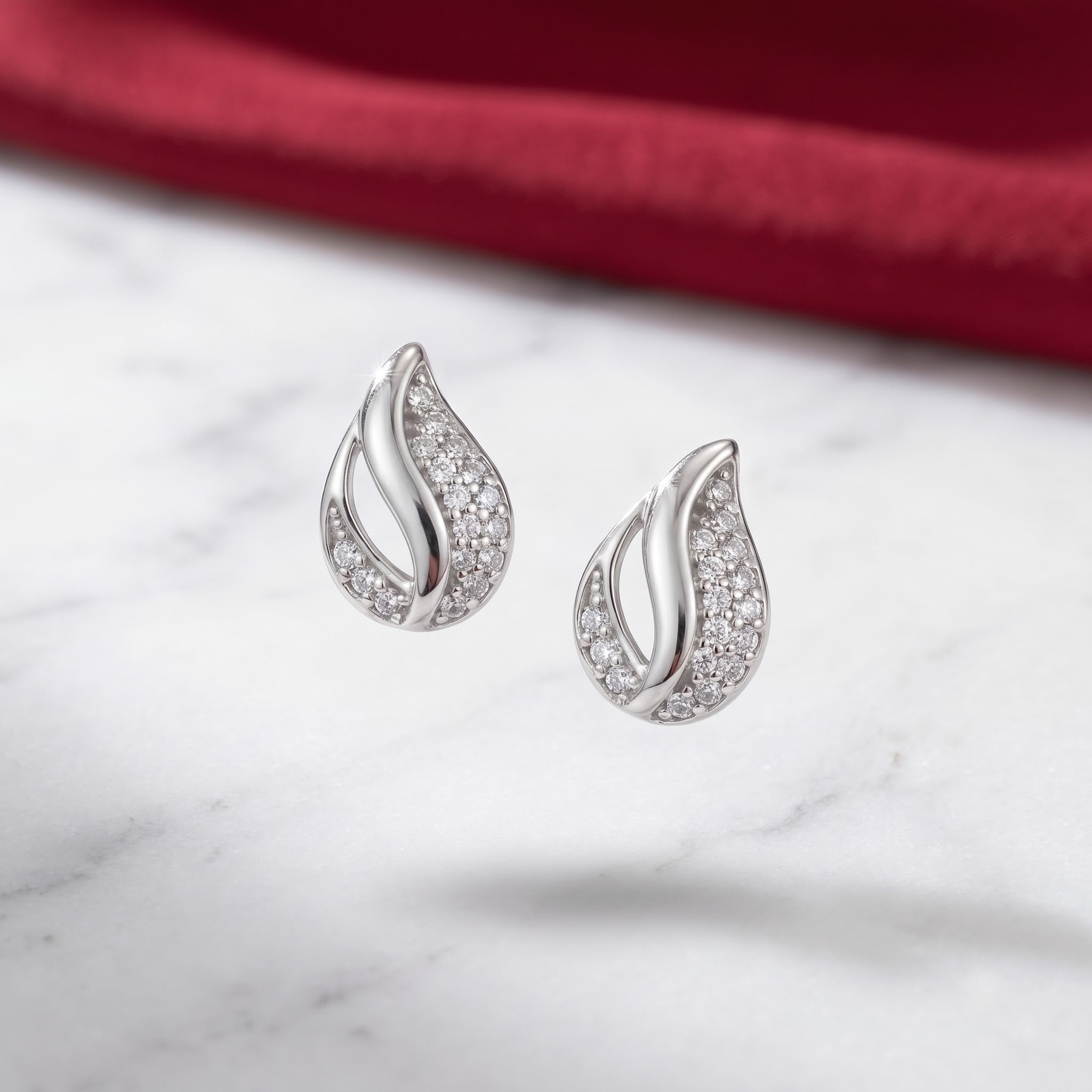 A pair of silver teardrop-shaped stud earrings paved with small clear stones, resting on a white marble surface with a blurred dark red fabric background.