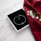A silver chain bracelet with a heart-shaped green gemstone centerpiece, displayed on a black jewelry box lid over a white marble surface with a red fabric and small white flowers.