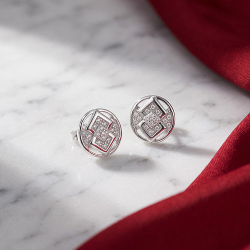 A pair of silver-toned geometric stud earrings with pavé-set clear stones, displayed on a white marble surface with a draped red fabric accent.