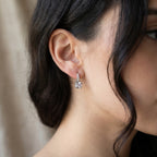 A close-up of a woman's ear wearing a small silver dangle earring with a clear faceted gemstone.
