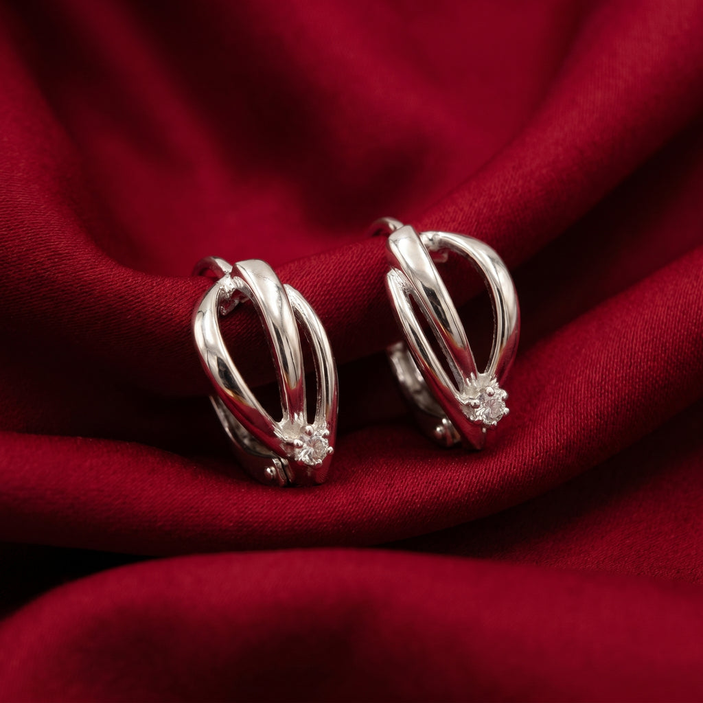 A pair of silver-toned huggie hoop earrings with a small clear crystal accent, displayed on draped red fabric.