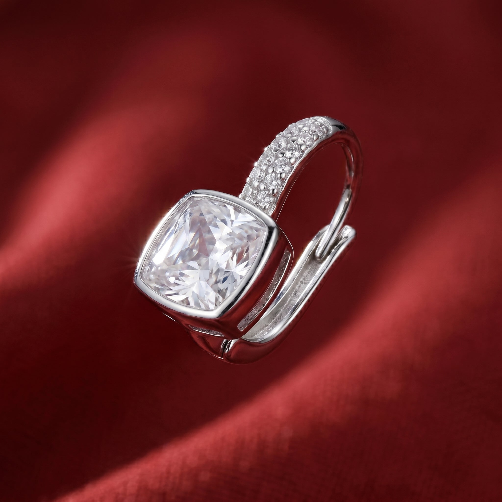 A silver-toned ring featuring a large cushion-cut clear gemstone and a pavé-set band, displayed on a draped dark red fabric background.