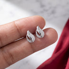 A pair of silver leaf-shaped stud earrings with pavé-set clear stones, shown resting on a person's fingers against a light marble background with a red fabric accent.