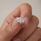 A close-up of a hand holding a round silver ring with a geometric diamond-pattern center.