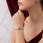 A close-up lifestyle shot of a model wearing a silver-toned heart-shaped bracelet with a green heart-shaped emerald and a row of clear stones, paired with a deep red velvet top.