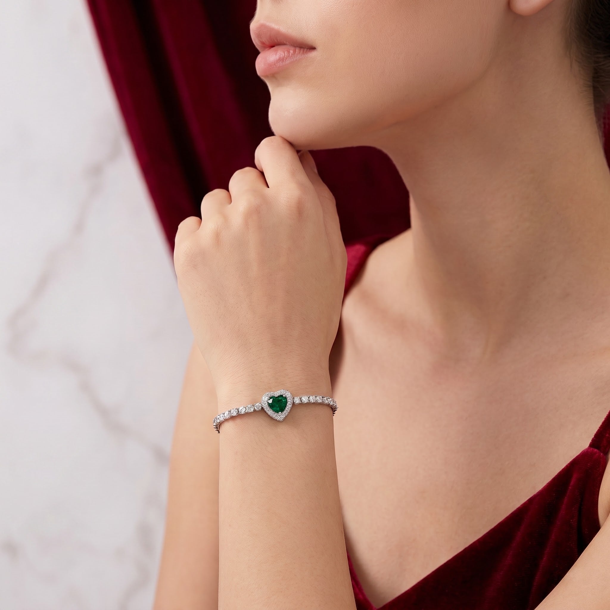 A close-up lifestyle shot of a model wearing a silver-toned heart-shaped bracelet with a green heart-shaped emerald and a row of clear stones, paired with a deep red velvet top.