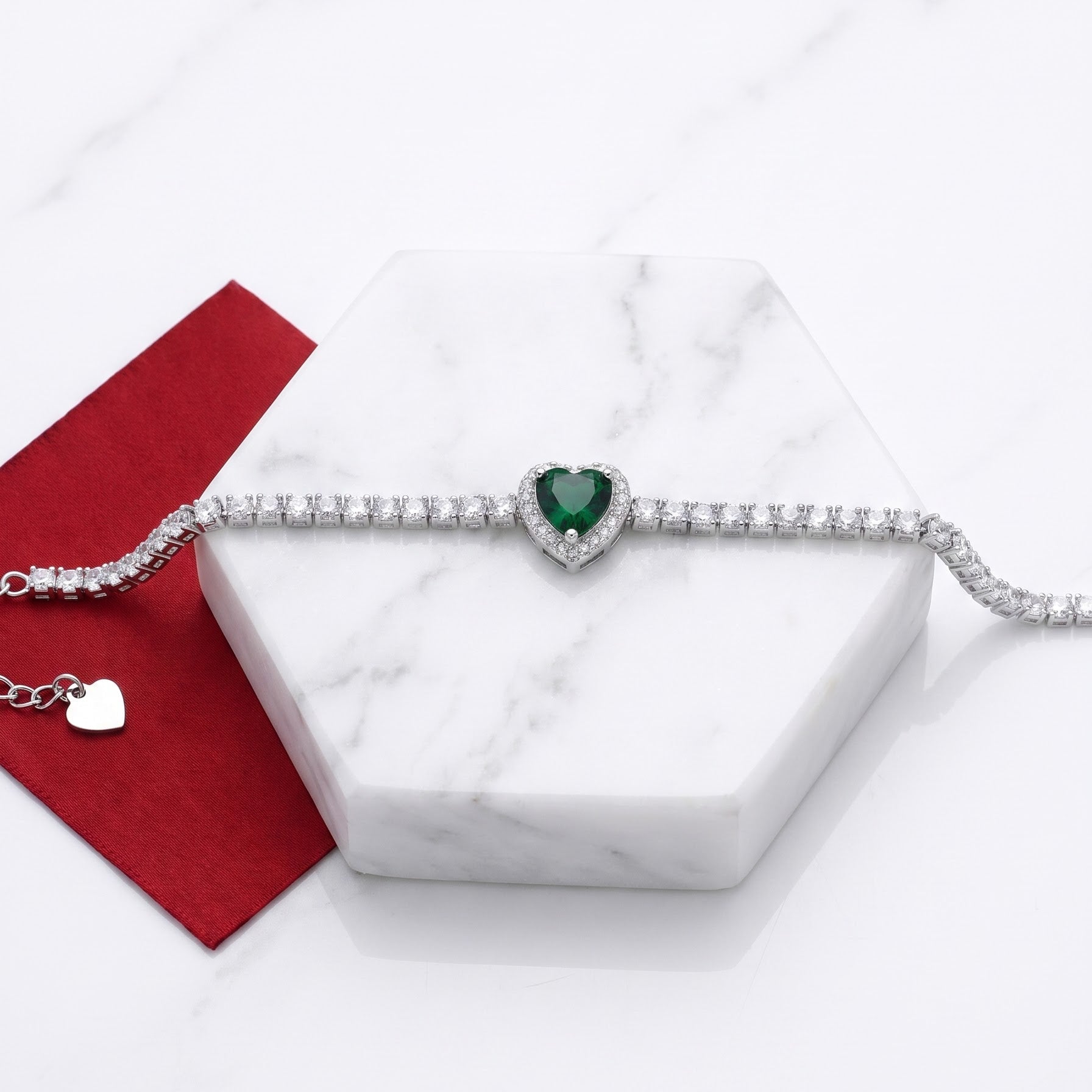 A silver-toned heart bracelet with a central green heart gemstone, displayed on a white marble hexagonal block next to a red velvet ribbon and a small silver heart charm.